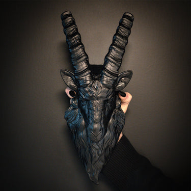 Black goat mask with large horns held against a dark background the blackened teeth