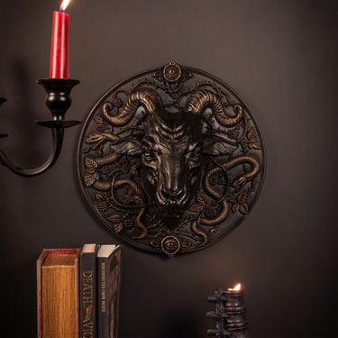 A circular resin gothic wall plaque featuring a detailed ram's head surrounded by coiled snakes, styled on a dark wall with a candle holder, books, and another candle.