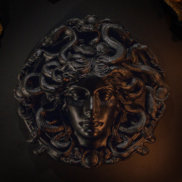 A dark metallic wall medusa gothic sculpture of a classical female face with snakes winding through her hair, shown against a dark background.