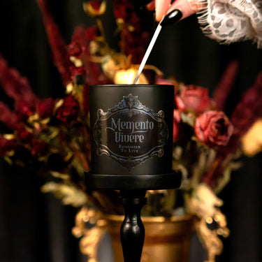 A black scented candle in a black holder with a decorative silver crest label reading 'Memento Vivere Remember To Live'. A hand with black nail polish inserts a wick into the candle, set against a dark background with blurred red and gold dried flowers.