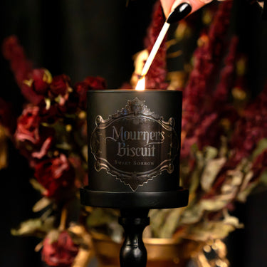 A black scented candle labeled 'Mourmers Biscuit Sweet Norrow' on a black candlestick holder, lit with a match against a dark background with red dried flowers.