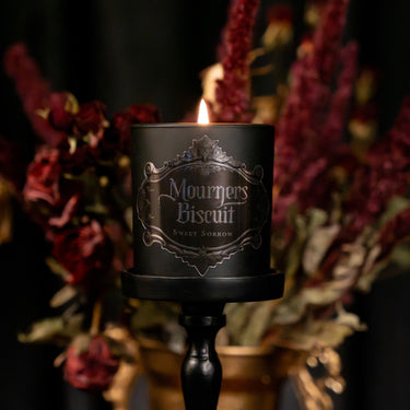 A black candle in a matte cylindrical holder on a black stand, lit with a flame. The front label reads 'Mourners Biscuit' and 'Sweet Sorrow' in ornate silver lettering.