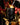 A black candle labeled 'Spiced Pumpkin Limited Edition' on a black candlestick holder, lit with a feathered lighter against a dark, blurred background of red flowers.