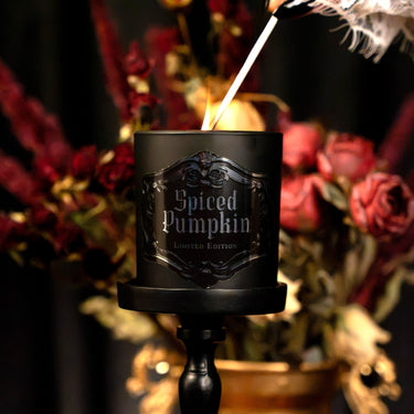 A black candle labeled 'Spiced Pumpkin Limited Edition' on a black candlestick holder, lit with a feathered lighter against a dark, blurred background of red flowers.