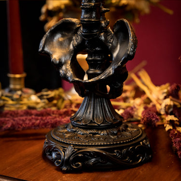 An ornate black metal candle holder with a sculptural, winged base and decorative scrollwork, displayed on a wooden surface with dried flowers and a red background. dark romantic gothic home interior