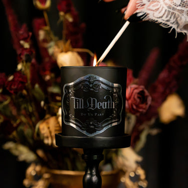 A black candle jar on a black candle holder, being lit with a match. The jar features a metallic silver gothic-style label that reads 'Till Death Do Us Part'.